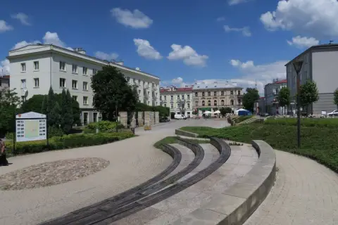 Słoneczny dzień na placu artystów w Kielcach. Widać budynki, zieleń i ławki.