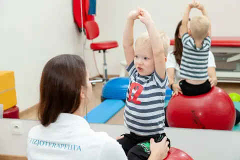 Fizjoterapeutka wspiera chłopca z zaburzeniami rozwojowymi podczas ćwiczeń na piłce rehabilitacyjnej.