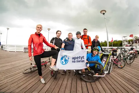 Grupa rowerzystów z banerem "apetyt na świat" pozuje do zdjęcia na molo, gotowi na wyprawę rowerem na Hel.