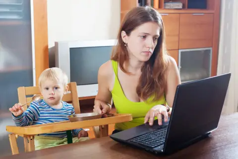 Mama na zasiłku macierzyńskim pracuje przy laptopie, podczas gdy jej dziecko siedzi w krzesełku i trzyma łyżeczkę.
