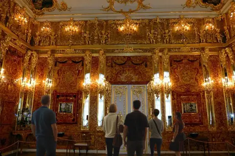 Zachwycająca bursztynowa komnata, gdzie złote zdobienia i światło tworzą magiczną atmosferę. Zwiedzający podziwiają jej piękno.