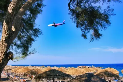 Samolot przelatuje nad plażą z parasolami, tuż przed lądowaniem na **Santorini lotnisko**.