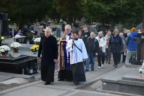 Ksiądz i ministrant niosą urnę na pogrzebie, a za nimi idzie żałobna procesja przez cmentarz.