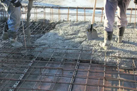 Betonowanie stropu. Robotnicy w gumowcach wylewają beton na zbrojenie. Łopata czeka na pomoc.