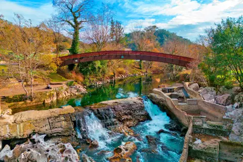 Cascada y puente de madera en la vía verde Puente Viesgo, rodeados de árboles otoñales y un cielo azul vibrante.