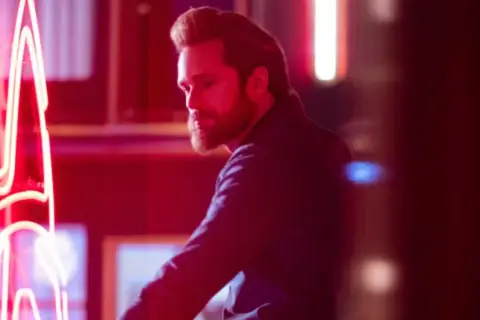 Mężczyzna z brodą w ciemnej koszuli odwraca się od neonowego napisu. Scena z programu telewizyjnego "Bastion".