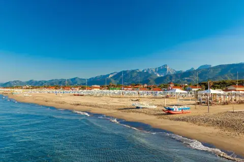 Słoneczna plaża w Toskanii, z rzędami białych parasoli i leżaków, w tle góry i błękitne morze.