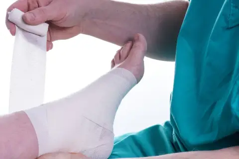 Opatrywanie stopy po kontuzji. Ręce owijają bandażem kostkę i stopę.