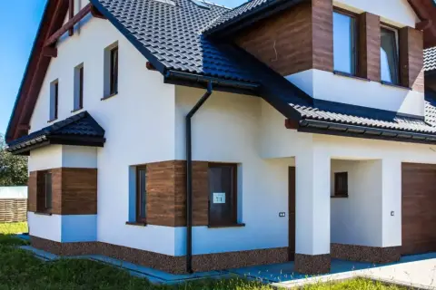 Nowoczesny dom z białym tynkiem i drewnianymi akcentami. Widoczny ciemny dach, okna i drzwi. Na dole domu ozdobny cokół.