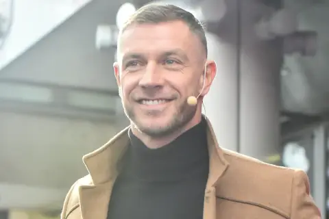 Uśmiechnięty Dariusz Pachut w beżowym płaszczu i czarnym golfie, z mikrofonem przy uchu. Może opowiada o swojej żonie.