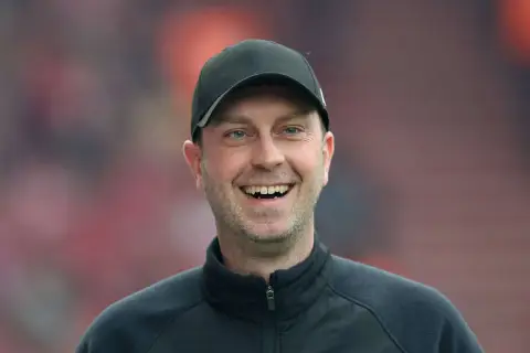 Lachender rb leipzig trainer mit schwarzer Kappe und Trainingsjacke vor unscharfem Publikum.