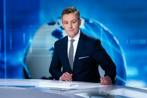 Elegancki konferansjer w garniturze stoi przy biurku na tle niebieskiej grafiki z globusem.