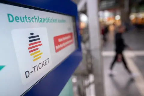 Das Deutschlandticket-Logo auf einem Bildschirm. Eine Person geht unscharf im Hintergrund vorbei. Vielleicht kann man das Deutschlandticket pausieren.
