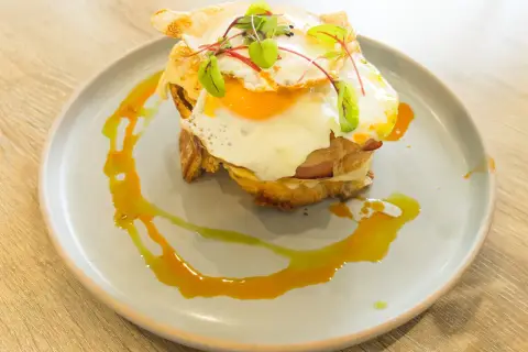 Un délicieux croque madame avec un œuf au plat coulant, du jambon et du fromage fondu, garni de jeunes pousses et servi sur une assiette avec un filet de sauce.