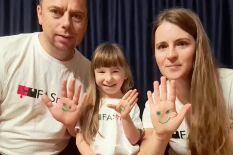 Uśmiechnięci dorośli z FAS i ich córka pokazują dłonie z namalowanymi buźkami.