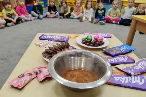 Dzieci w przedszkolu świętują dzień czekolady. Na stole czekolady Milka, Kinder, ciastka i miska z kakao.