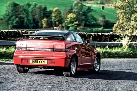 Czerwone Alfa Romeo SZ z czarnym dachem i spoilerem jedzie po wiejskiej drodze.