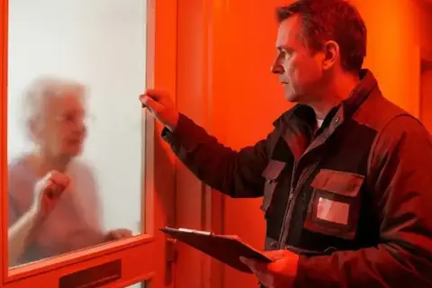 Mężczyzna puka do drzwi starszej pani, trzymając dokumenty. Uważaj na oszustów podszywających się pod pracowników Tauron, którzy chcą wymienić licznik.