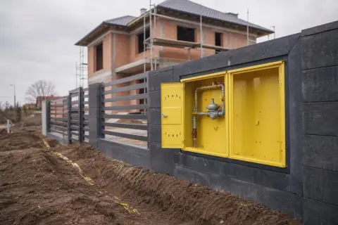 Żółta skrzynka z instalacją gazową, widoczna na tle budowanego domu i ogrodzenia.