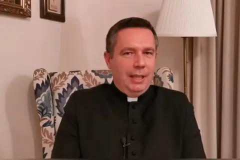 Ksiądz w czarnej sutannie siedzi w fotelu. Mówi o tym, ile za mszę w niedzielę, a za nim widać lampę i obrazy.
