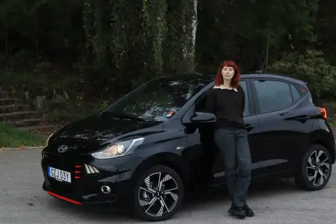 En kvinna med rött hår lutar sig mot en svart Hyundai i10. Bilen är redo för ett test.