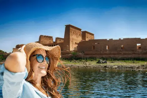 Mujer con sombrero y gafas de sol sonríe en un barco, con templos egipcios de fondo. ¡La mejor época para viajar a Egipto es ahora!