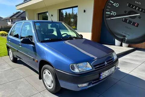 Niebieski Citroen Saxo z niskim przebiegiem, zaparkowany przed nowoczesnym domem. Na desce rozdzielczej widać licznik z przebiegiem 14649 km.