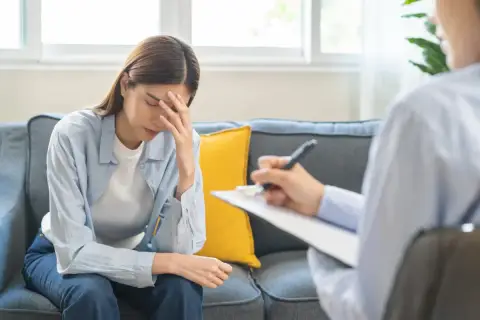 Kobieta z ręką na czole siedzi na kanapie, rozmawiając z terapeutą. Może to być rozmowa o diagnozie Aspergera.