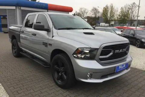 Srebrny Dodge Ram 1500 5.7 Hemi z czarnymi felgami i przyciemnianymi szybami stoi na parkingu przed budynkiem z niebiesko-pomarańczowym banerem.