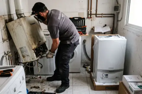 Técnico trabajando para cambiar caldera de gas. Retira una vieja y sucia caldera Junkers, mientras una nueva Vaillant espera en su caja.
