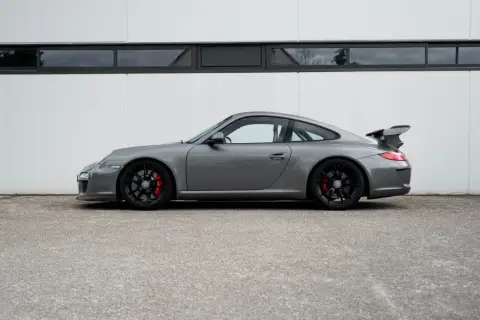 Grauer Porsche 997 GT3 RS mit schwarzem Flügel und roten Bremssätteln steht vor einer weißen Wand.