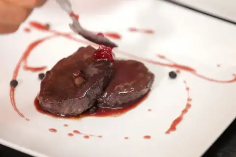 Elegancko podany comber z jelenia, polany sosem z owoców leśnych, ozdobiony ziarnami pieprzu i jagodami.