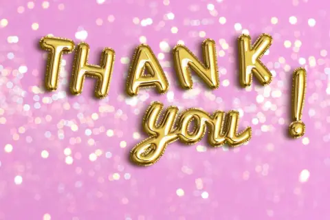 Globos dorados forman las palabras de agradecimiento "Thank you!" sobre un fondo rosa con luces bokeh.