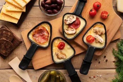 Raclette party with melted cheese, bacon, mushrooms, and tomatoes. Discover par quoi remplacer le fromage à raclette pour varier les plaisirs !