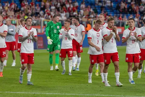 Piłkarze w białych strojach z orzełkiem na piersi, być może przyszli mistrzowie polski w piłce nożnej, stoją na murawie.