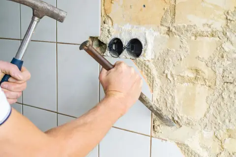 Ręka z młotkiem i dłutem usuwa płytki z płyty gipsowej, odsłaniając puste puszki elektryczne.