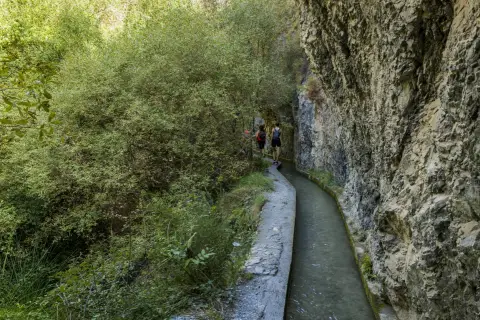 Senderistas caminan por la ruta de los bolos, un sendero estrecho junto a un canal de agua y un acantilado rocoso.