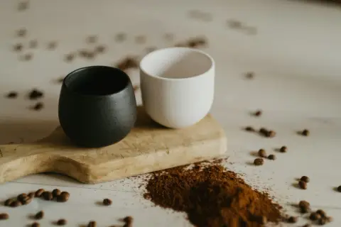 Dwa ceramiczne kubki, czarny i biały, na drewnianej desce. Obok rozsypana mielona kawa, która sugeruje, czy kawe sypana sie miesza.