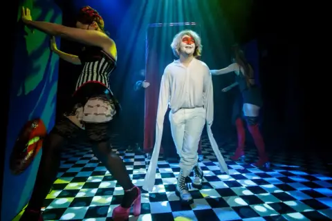 Scena teatralna, gdzie młody mężczyzna w białym stroju i makijażu twarzy stoi pośród postaci w kostiumach, tworząc atmosferę tragifarsy.