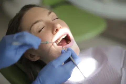 Kobieta z zamkniętymi oczami poddaje się zabiegowi stomatologicznemu. Dentysta w niebieskich rękawiczkach wykonuje kiretaż zamknięty, używając narzędzi do oczyszczania zębów.