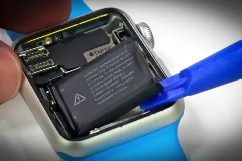 Wymiana baterii Apple Watch: wyjmowanie starego ogniwa za pomocą niebieskiego narzędzia.