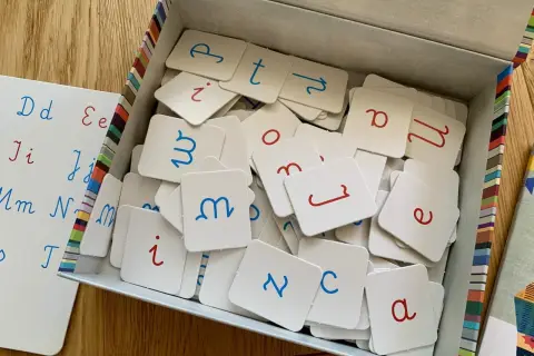 Pudełko z kartami do nauki polskiego alfabetu pisanego. Litery w kolorze niebieskim i czerwonym. Obok biała tablica z zapisanymi literami.