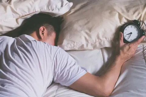 Mann schläft im Bett, Wecker liegt daneben. Wie viel Schlaf braucht man wirklich?
