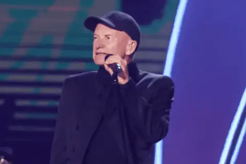 Sting w czapce i czarnym stroju śpiewa do mikrofonu. Ciekawe, ile Sting bierze za koncert, gdy występuje na tak dużej scenie.