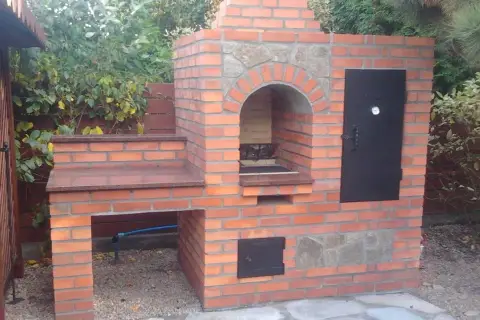 Solidny grill z cegły szamotowej, z kamiennym łukiem nad paleniskiem i metalowymi drzwiczkami. Idealny do ogrodu.