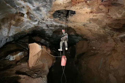 Odważny speleolog wspinający się po linie w jaskini, z workiem na sprzęt zwisającym poniżej.