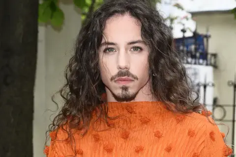 Michał Szpak w pomarańczowej bluzce z falbanami, z burzą loków i charakterystyczną bródką. Jego wiek nie wpływa na jego styl.