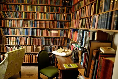 Wnętrze biblioteki z regałami pełnymi książek. Na biurku leży poduszka z książkami, a obok krzesło. Nawet niewierzący doceniłby bogactwo wiedzy.
