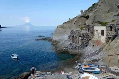 Sur l'île Salina, un homme amarre un bateau près de vieilles maisons de pêcheurs en pierre, avec une île volcanique au loin.