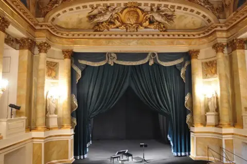 Złote zdobienia i bogate zasłony tworzą majestatyczne oświetlenie sceny teatralnej. Na scenie dwa stołki i pulpit nutowy czekają na artystów.